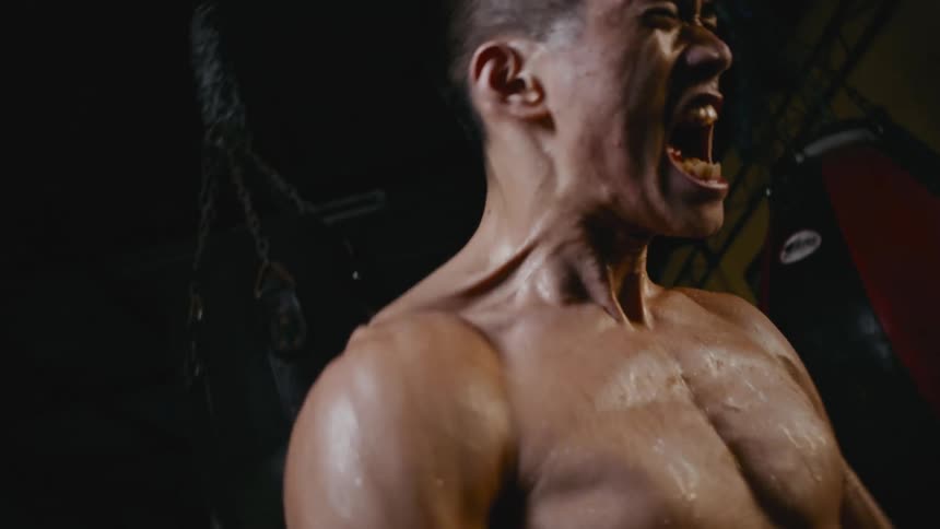 A man in boxing gloves stands in a gym and clenches his fists and yells