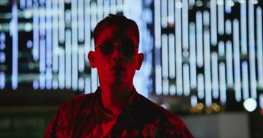 Young asian man wearing sunglasses standing in front of buildings in the city at night looks at the camera
