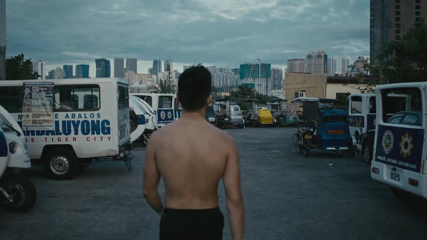 A shirtless man is walking outside while tall buildings can be seen at a distance
