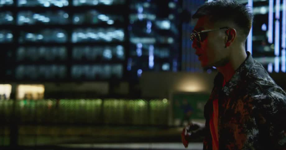 A man with sunglasses standing on a city street with a billboard and buildings in the background