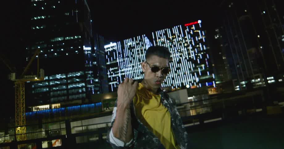 Dancer wearing sunglasses dancing in front of buildings in the city at night while looking at the camera