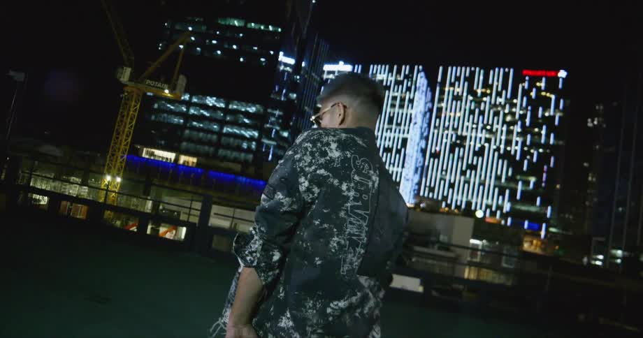 A man dances on a rooftop with lit up buildings behind him