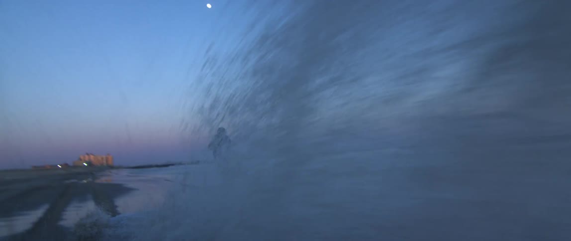 A man driving a mortocycle on the shoreline of a beach at sunset passes through a spray of water