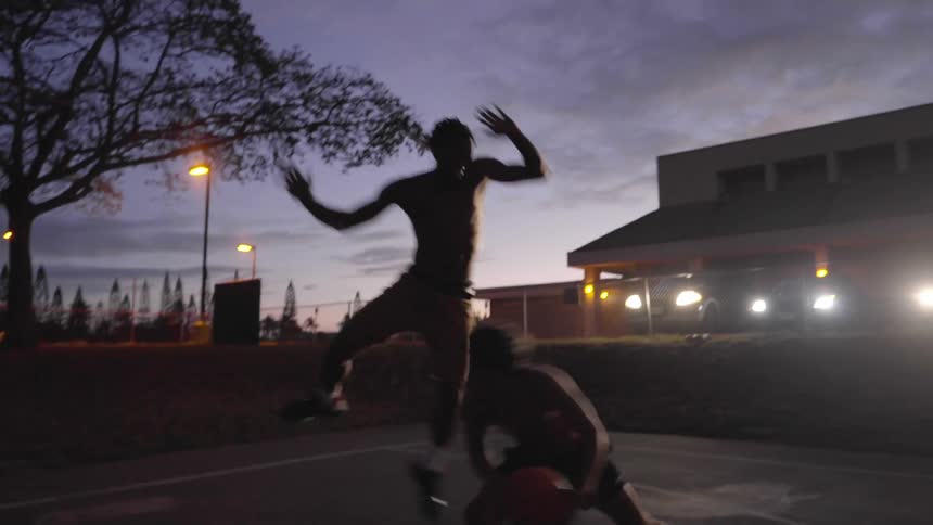 Shirtless two men play basketball in the basketball court at dusk