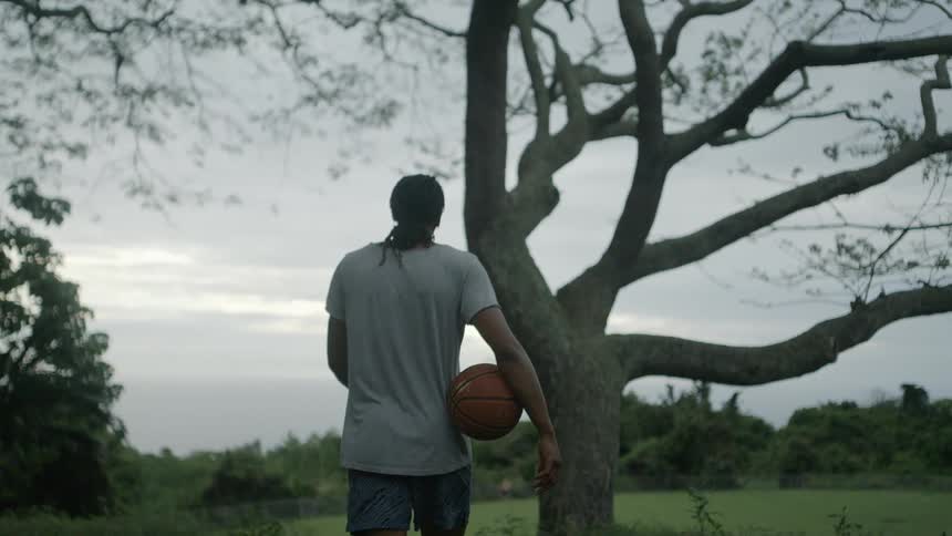Young man carrying a basketball walking near a tree in a field