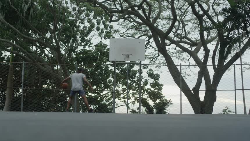A man on a basketball court shooting a basketball with trees the sky and clouds in the background