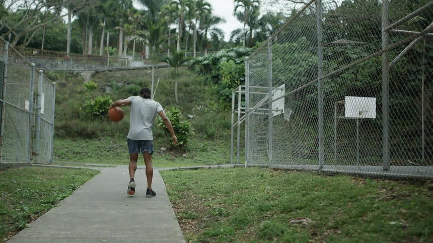 Man walks along sidewalk between next to fenced basketball court dribbling basketball