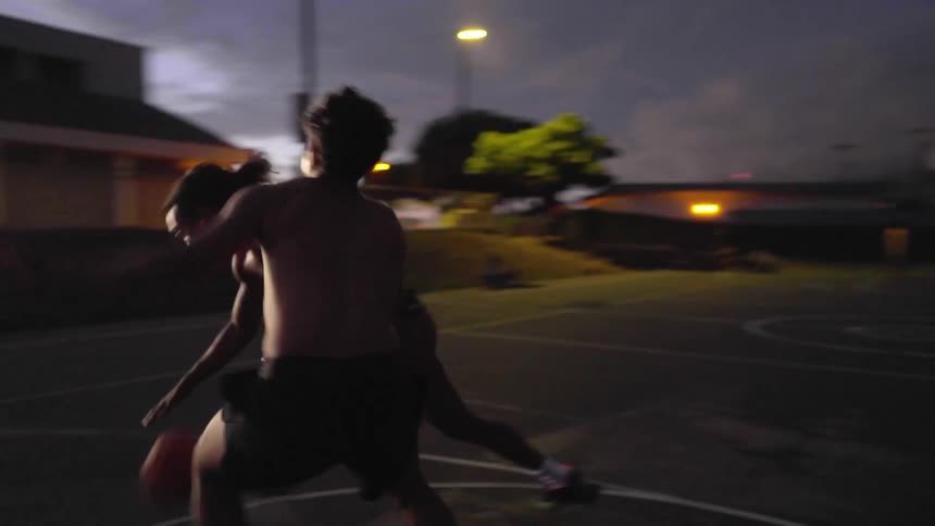 Shirtless two men play basketball in the basketball court at dusk