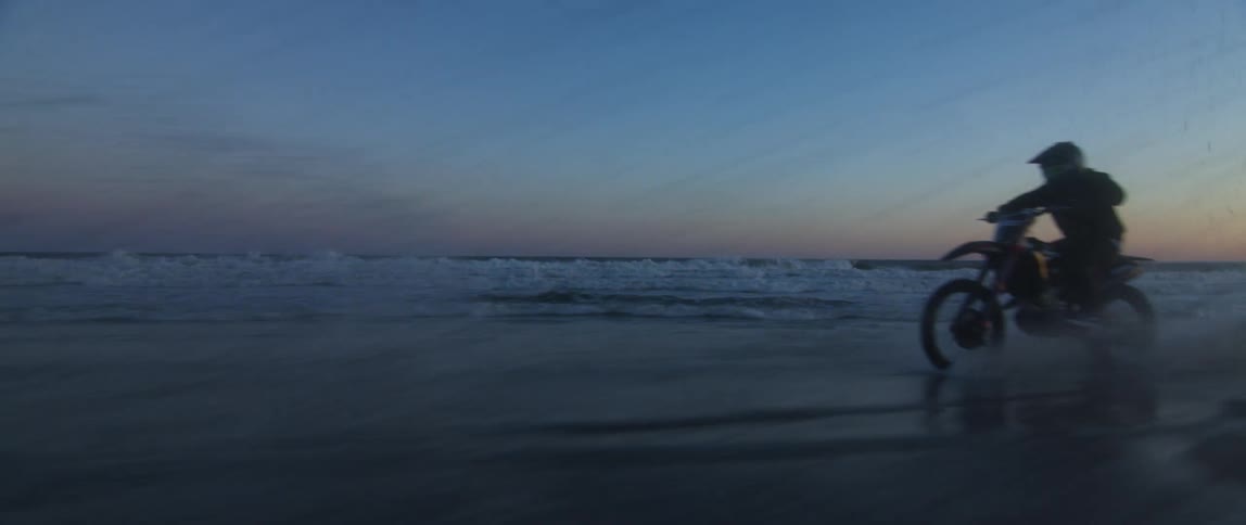 Male biker with a helmet riding bike along beach is silhouetted against the sky at dusk