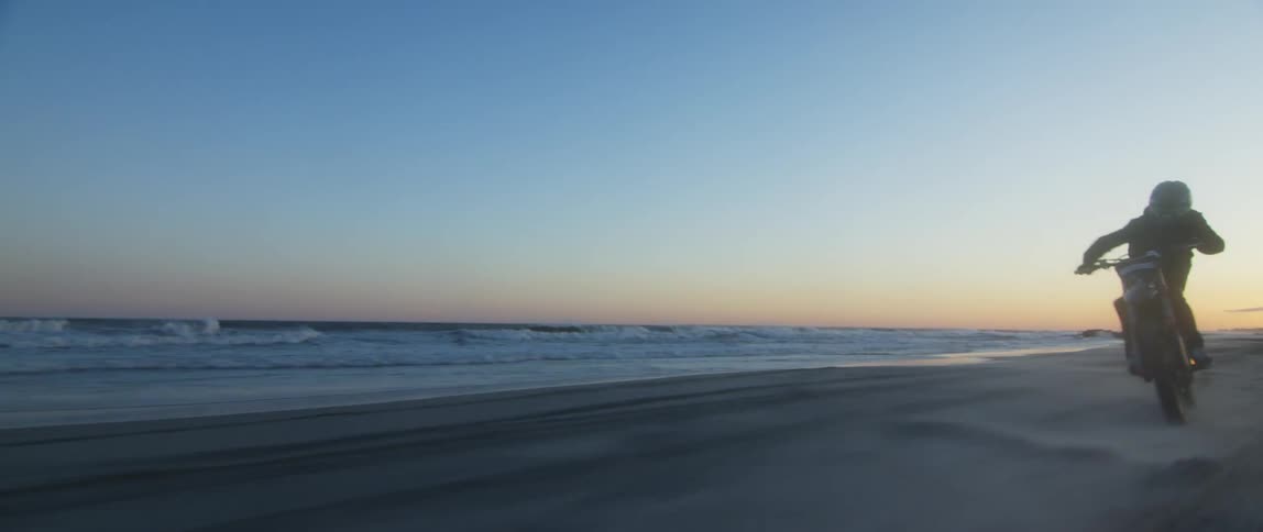 A person does a wheelie riding a motorbike on a beach with a low sun horizon in the distance