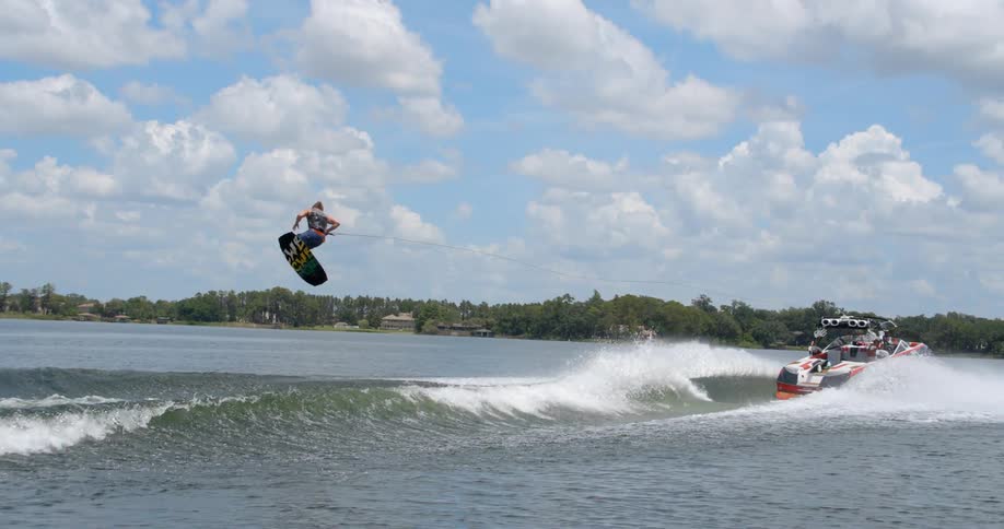 A man wakeboarding behind a boat spins in the air after being sent airborne by a wave before landing successfully
