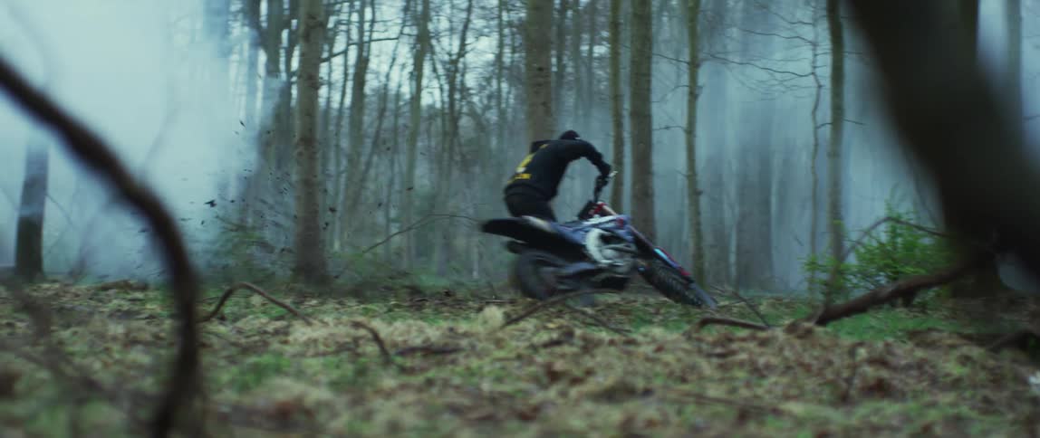 A man with a hat and hoodie revving a motorcycle in the mud in the forest with the sky and clouds in the background