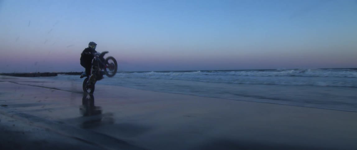 A man doing wheelies in a mortocycle on the shoreline of a beach at sunset