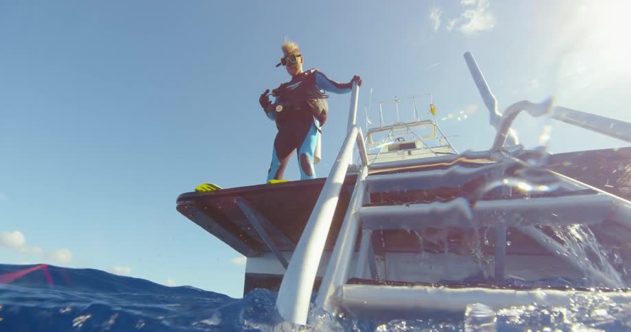 Blonde man wearing scuba gear stands at edge of boat bobbing in water under sunny lightly clouded sky