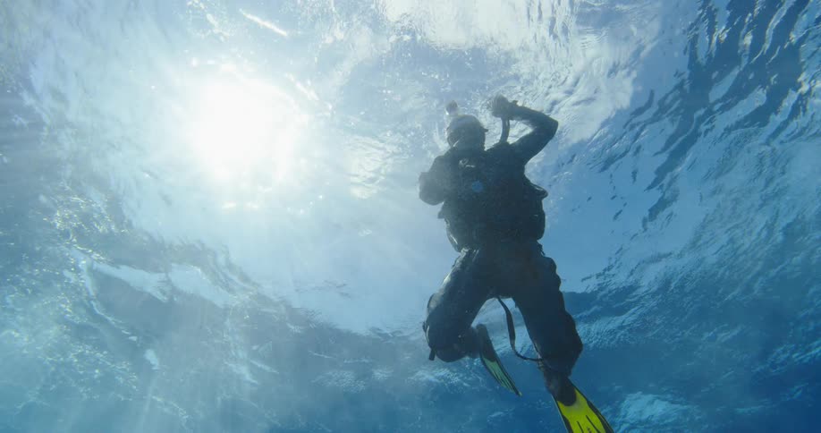 Scuba diver floats just beneath the surface blowing bubbles as the sun shines down through water from just over head