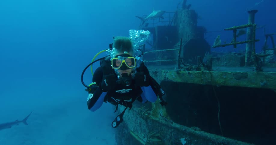 A man with scuba gear swimming underwater with sharks and a sunken ship in the background