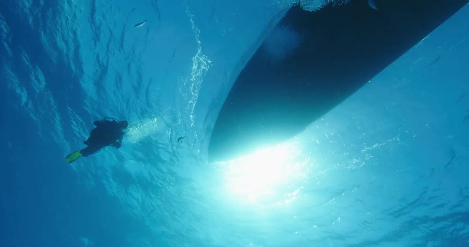 A person in scuba gear swimming underwater in the ocean with fishes and sharks next to a boat