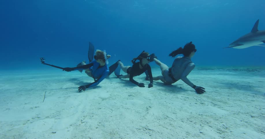 Trio of female freedivers swimming over the bottom of the ocean with sharks