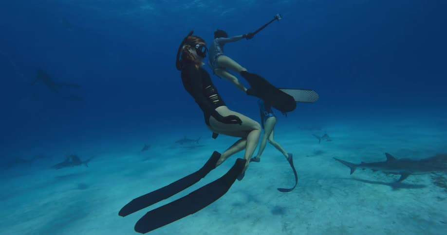 Three female divers swim slowly underwater as sharks swim close to seafloor