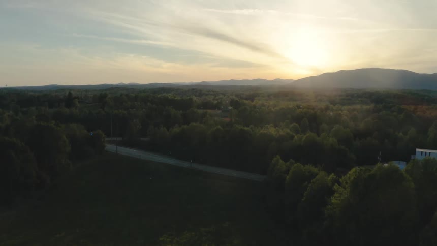 Road surrounded by many trees and the sun is above a mountain range in a cloudy sky