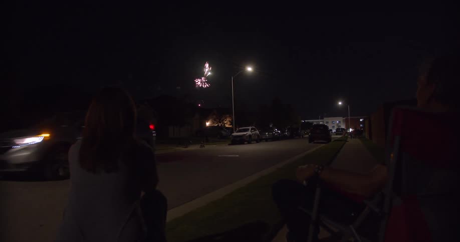 A woman stands on the side of the road and watches fireworks in the distance as a vehicle drives past