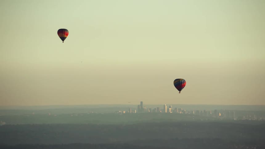 A pair of hot air balloons float over a forest outside of a metropolis during the day