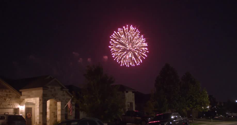 Fireworks display floats in sky above large houses with several cars parked on street