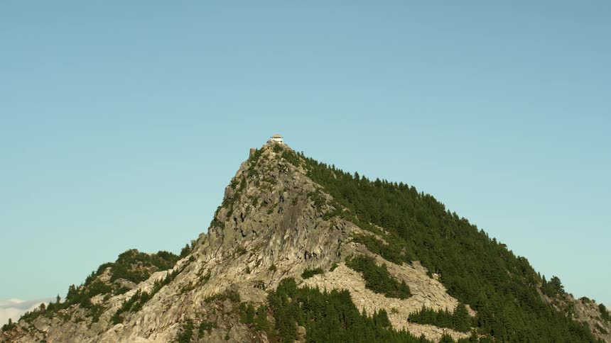 Building sits near peak of craggy forest covered mountain under a clear blue sky