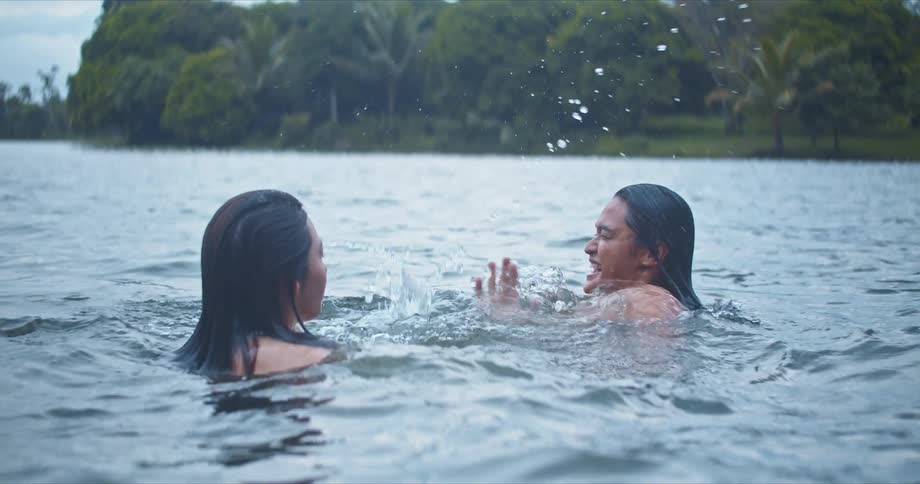 A man and a woman swimming in a lake with trees the sky and clouds in the background