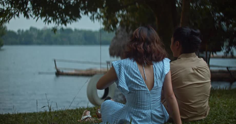 Two women sit in the grass and chat by a lake