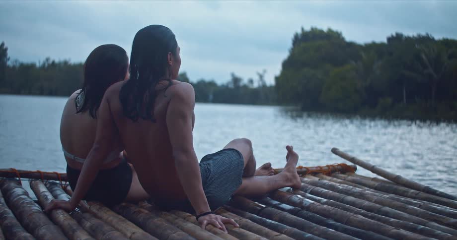 A man and woman sit side by side with legs outstretched on a rustic wooden raft