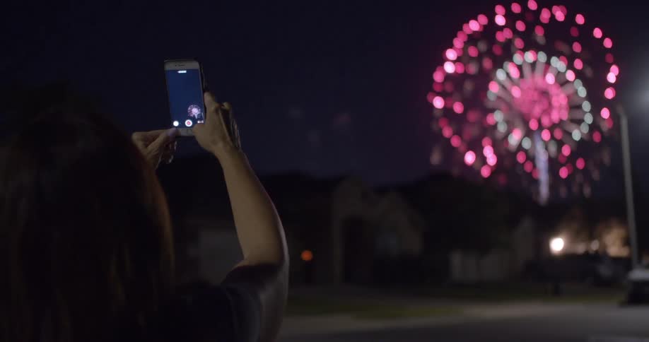 A woman stands outside and holds her cell phone up to take pictures of fireworks