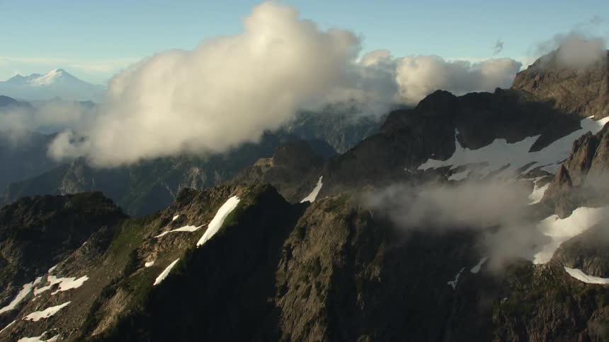 Clouds float over a barren mountain ranges covered in patches of snow on a sunny day