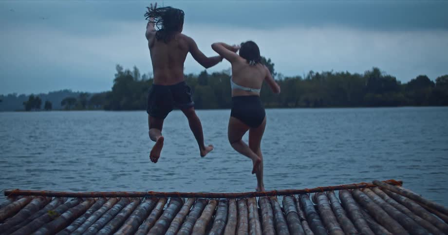 A man and a woman jumping from a pier into a lake with trees the sky and clouds in the background