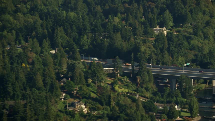 Vehicles driving on a bridge over a river with trees and buildings in the background
