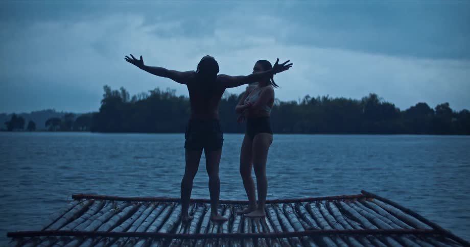 A man in swim trunks raises his face and arms towards the sky as he stands on a wooden pier with a woman in a swimsuit on a cloudy day