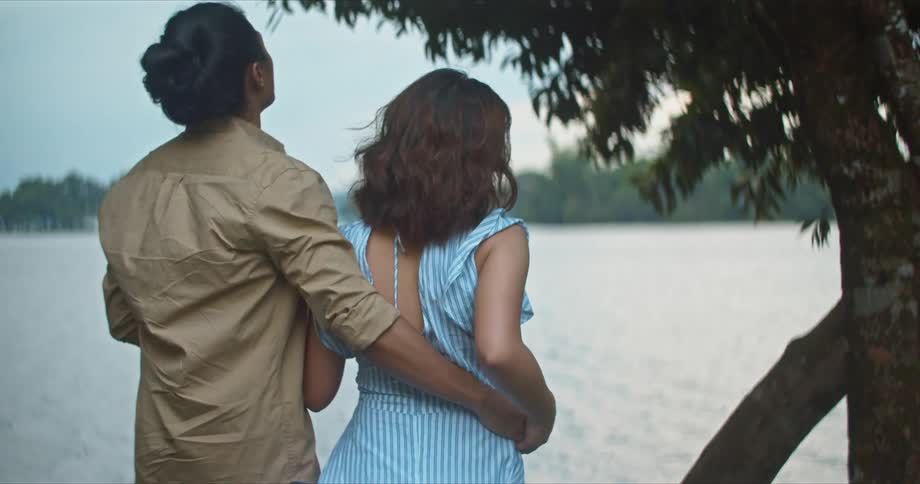 A man puts his arm around the waist of a woman as they stand together on the shore of a lake during the day