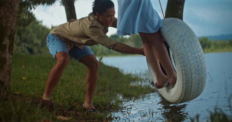 Woman wearing dress stands on tire swing being pushed by man over water