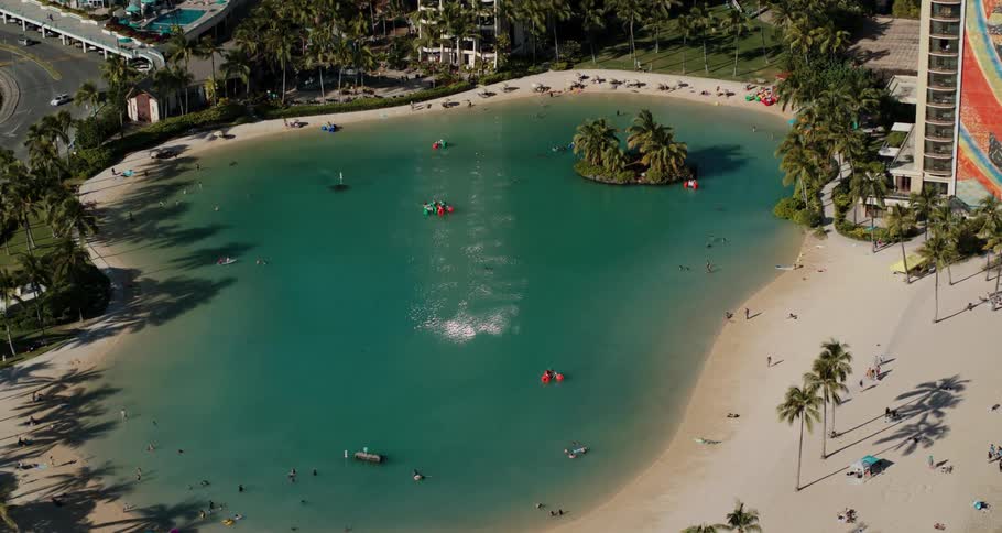 People move along sandy beach bordering green blue pond and buildings