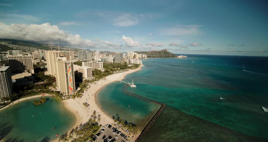 Palm trees dot light sandy beach separating city skyscrapers from deep green blue ocean through which vehicles move leaving white foam wakes