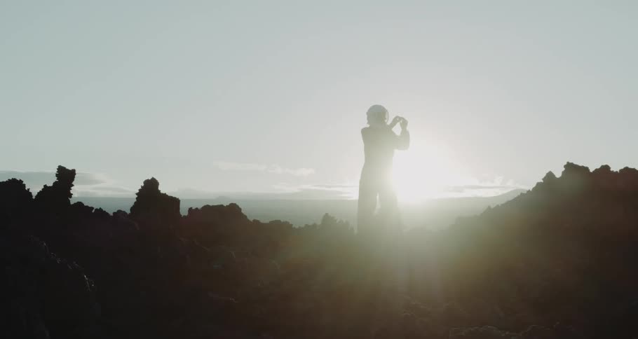 A person wearing an astronaut suit stands atop the rocks on a mountain top at sunset