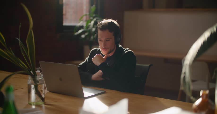 A man sitting at a table puts on a pair of headphones before typing on a laptop