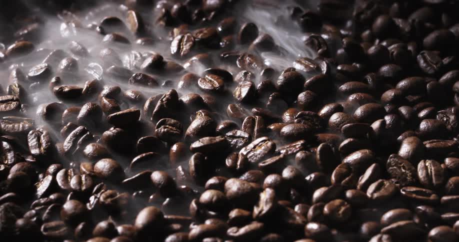 A layer of dark coffee beans release wisps of steam as they are roasted