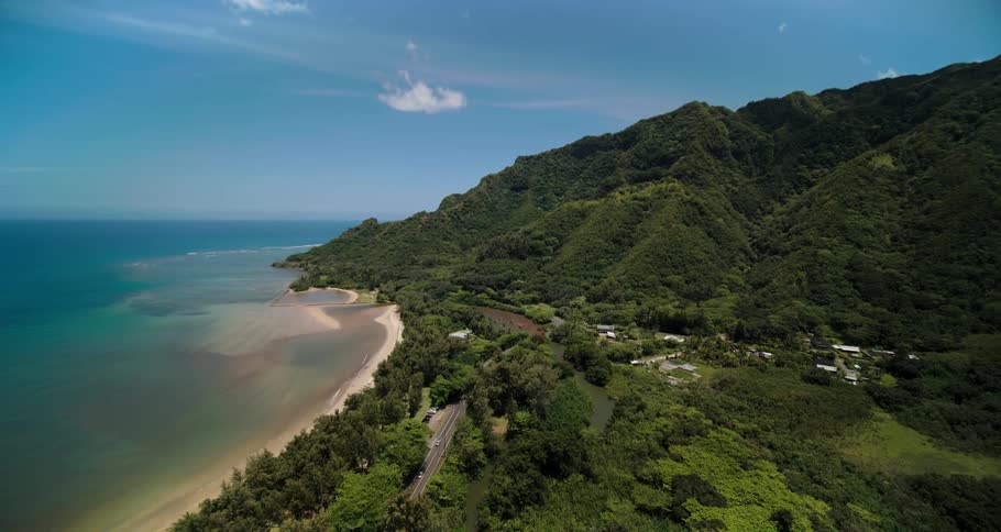 Traffic flows down road near beach bordered by trees leading to and from town towered over by forest covered town