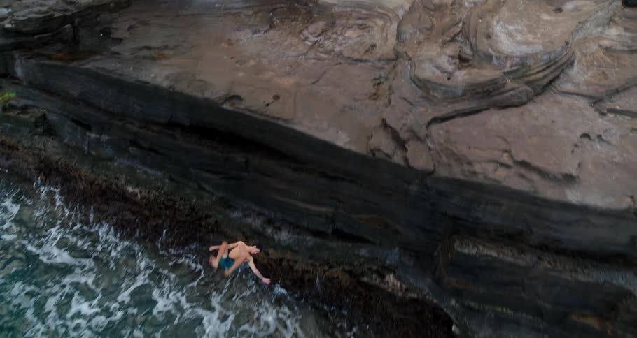 Man in shorts jumps off low cliff into blue ocean performing flip in midair