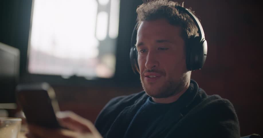 Man with a beard a black sweater and black headphones uses a smartphone