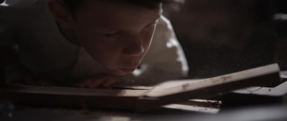 A boy blows saw dust off a piece of wood