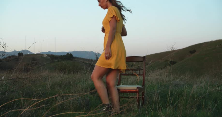 A model in a yellow sundress and combat boots walks through a field and sits down on a chair