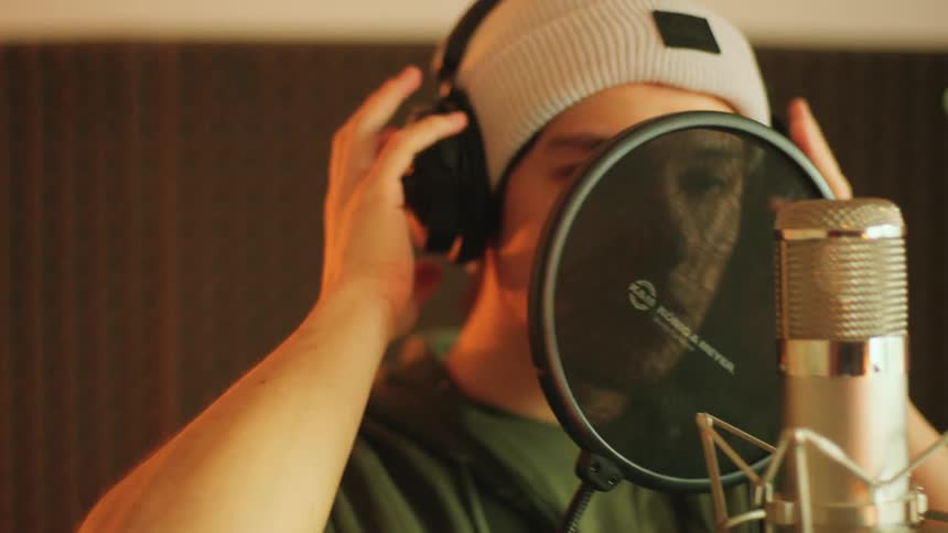 A man gestures with his hand as he sings into a recording microphone then takes off headphones