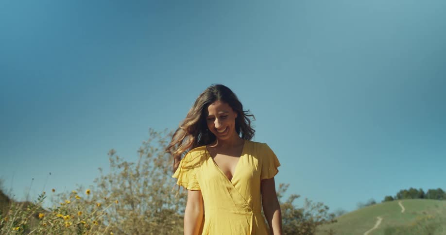 Woman wearing a yellow dress walking near a hill looks at the camera laughing